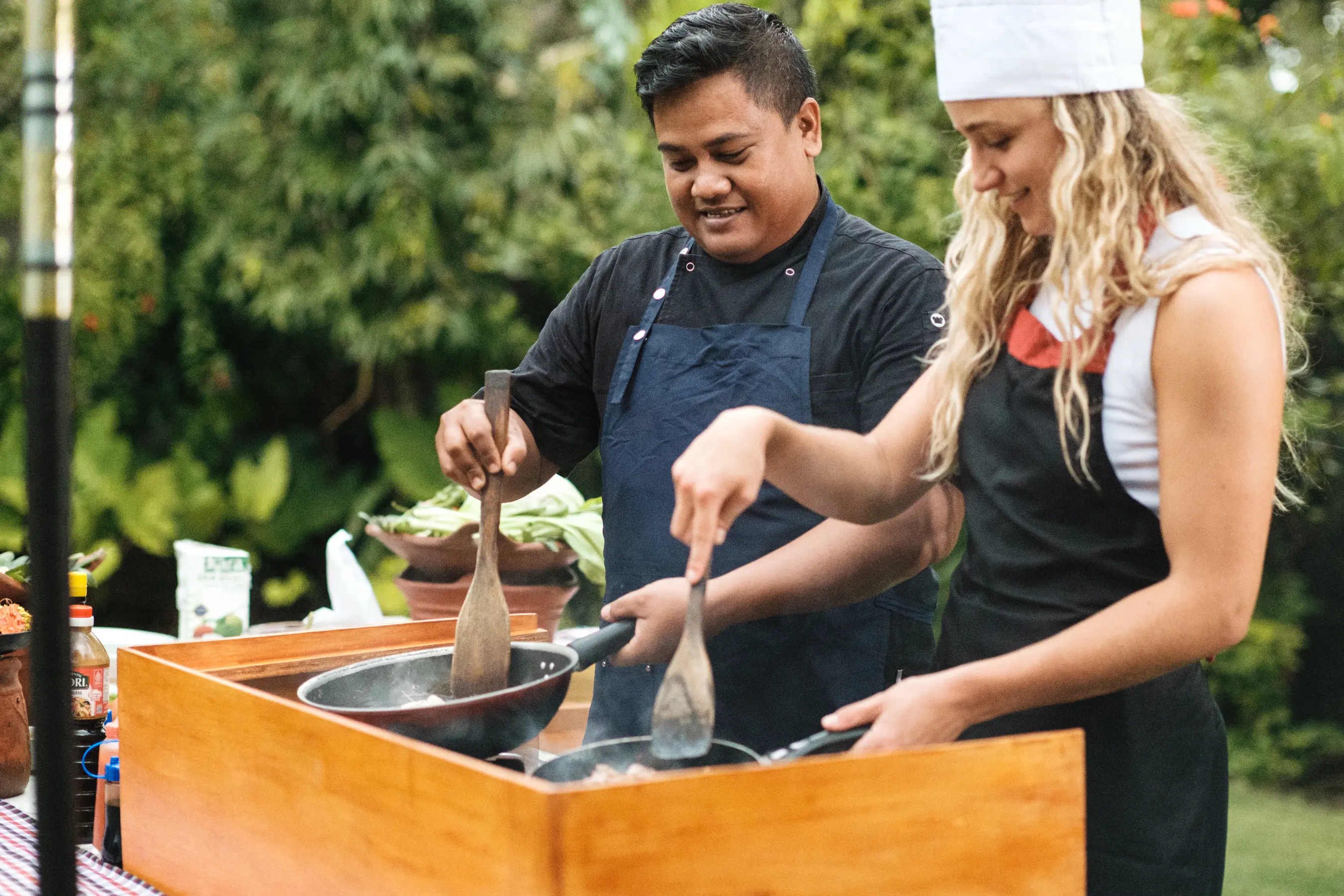 Two chefs cooking together at an outdoor wooden BBQ station during a private cooking class.