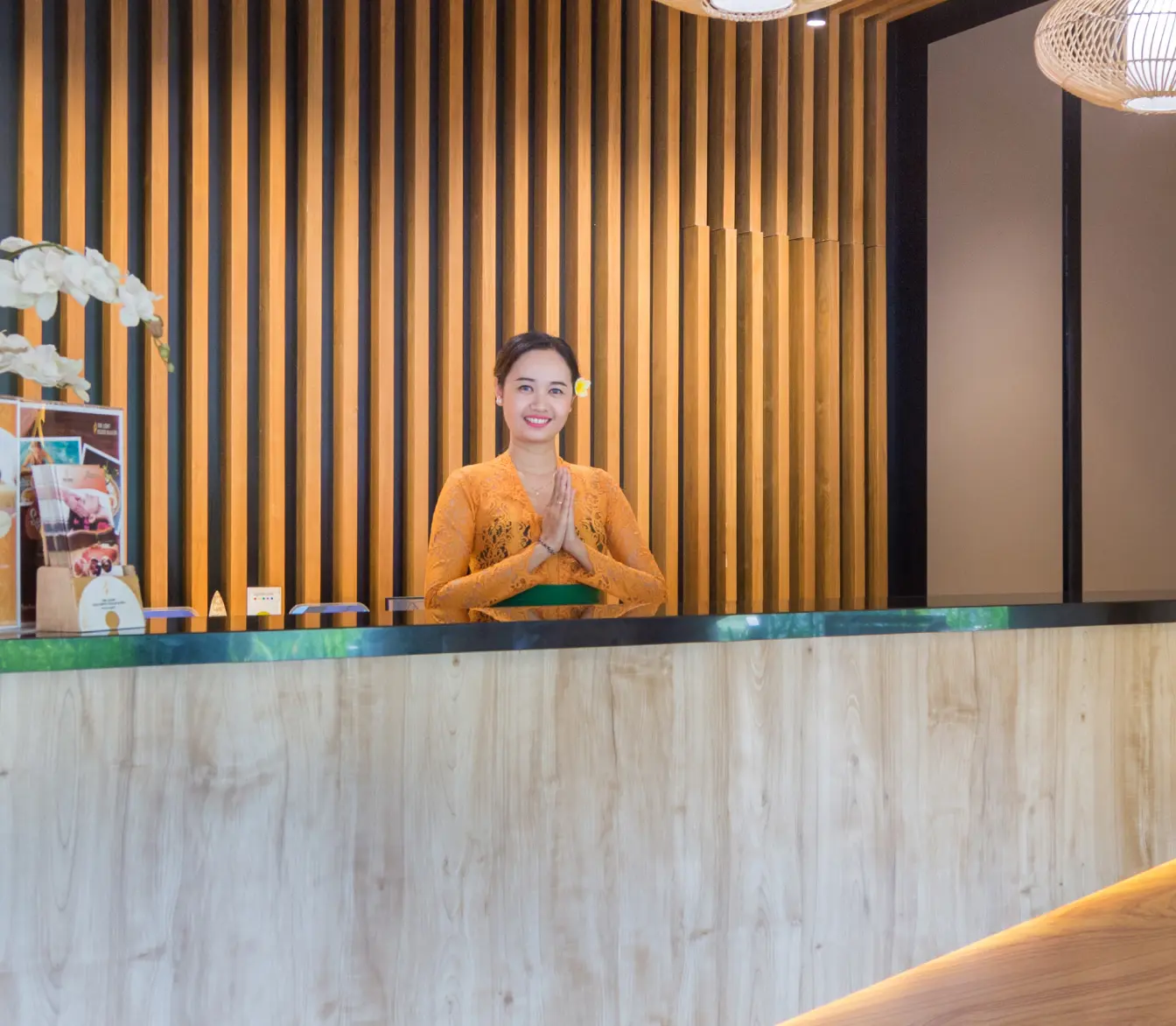 Female receptionist in traditional Balinese kebaya greeting with a sembah gesture behind a wooden reception desk.