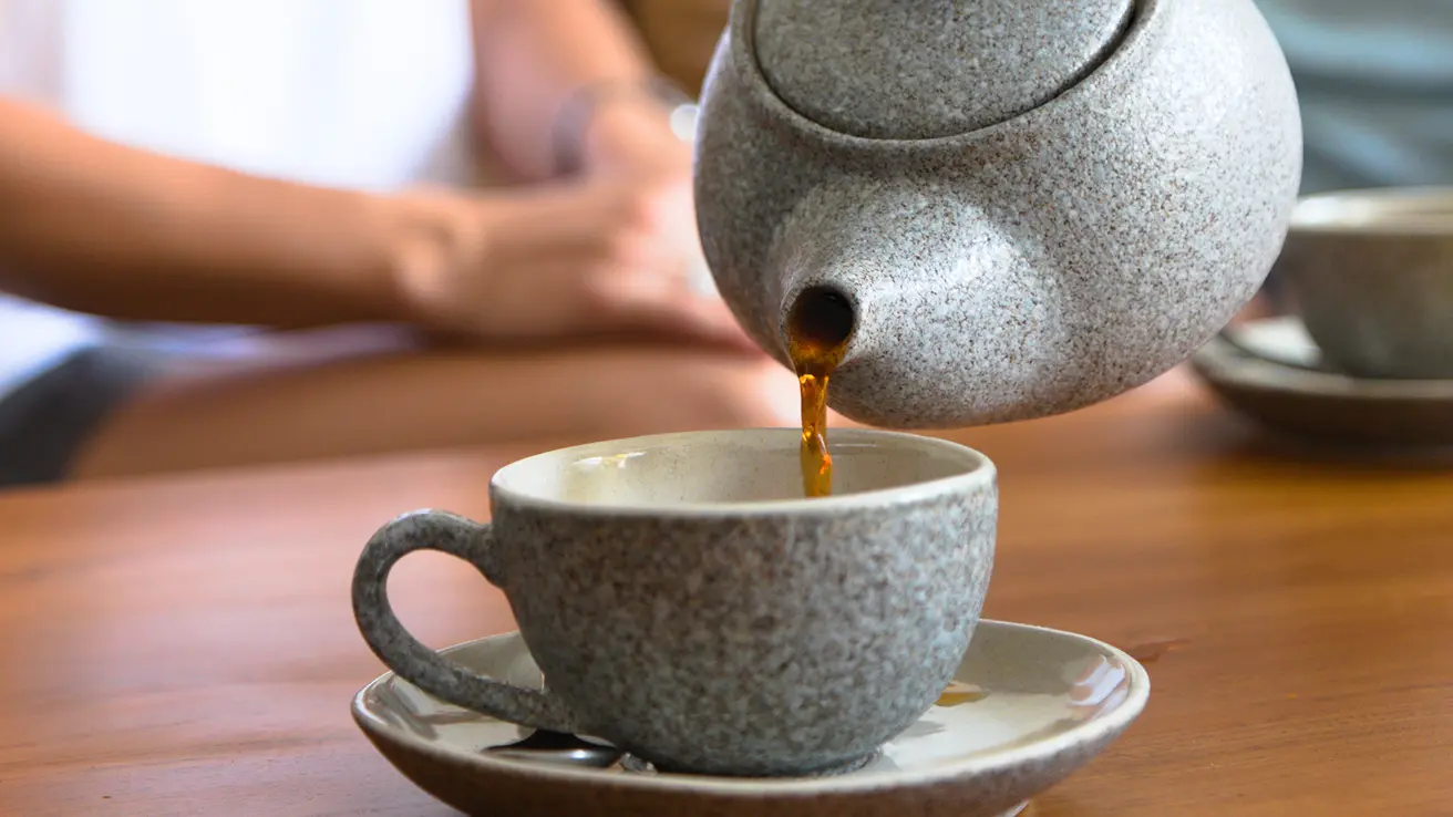 Close-up of tea being poured from a speckled stoneware teapot into a matching ceramic cup during afternoon tea service.