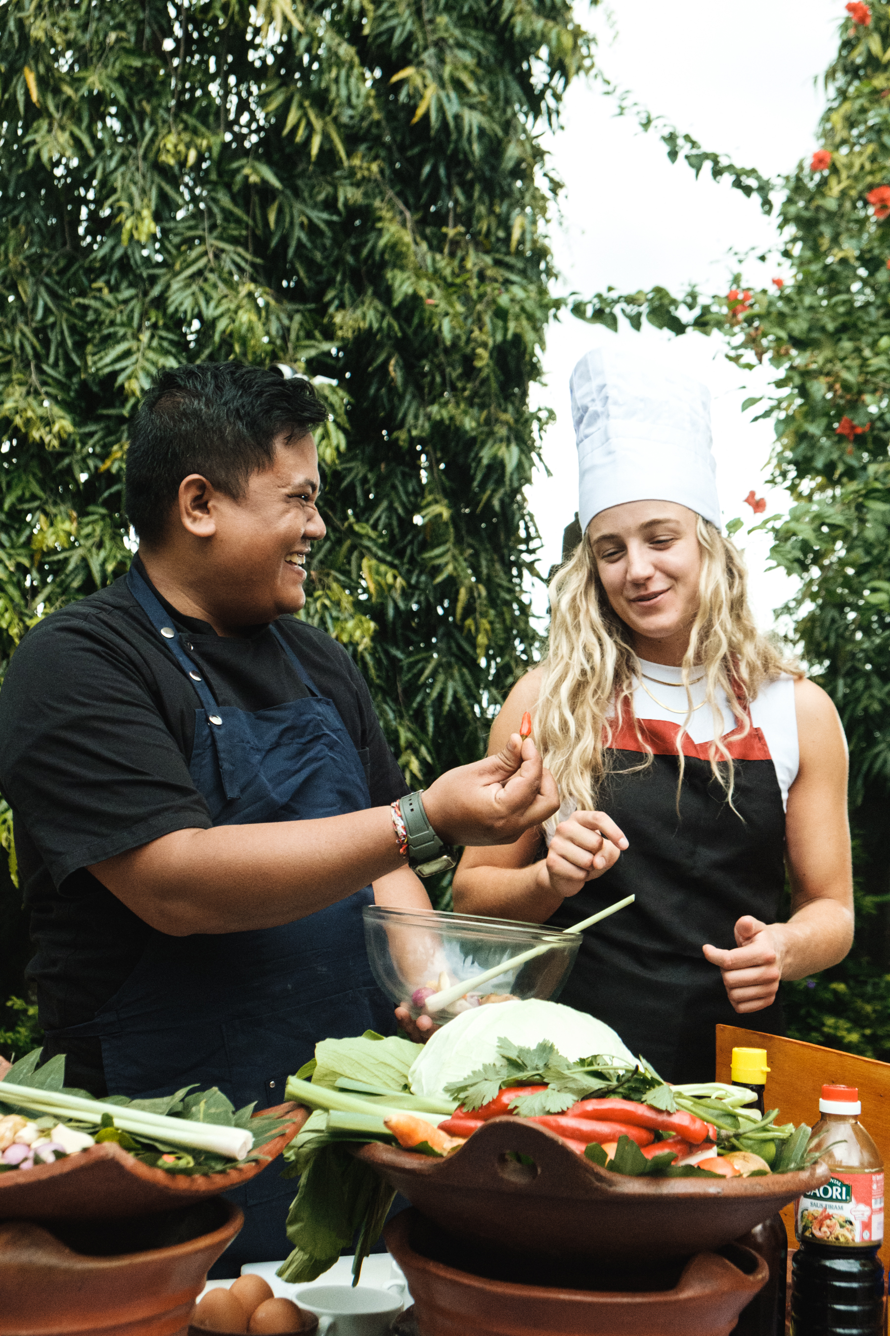 Local Balinese chef and a young woman in chef's hat laughing during an outdoor cooking with fresh vegetables.