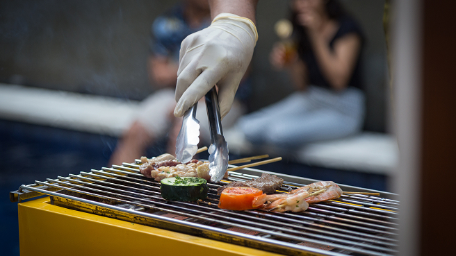 Gloved hand using metal tongs to grill skewers of prawns, beef, tomato and zucchini over a yellow charcoal BBQ.
