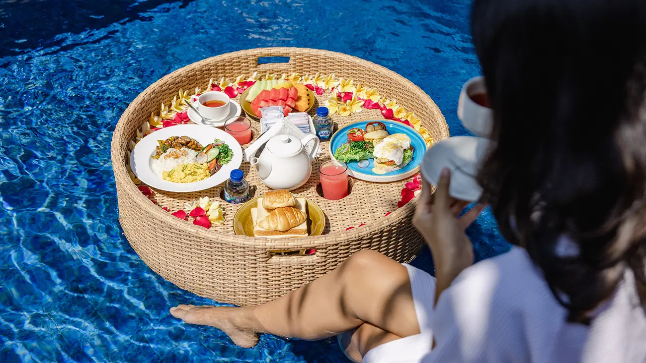 Woman in white robe enjoying a round wicker floating breakfast basket with eggs, fruit, juice and tea in a private pool.
