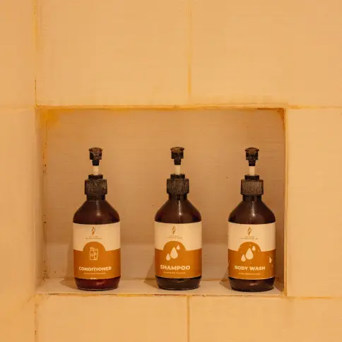 Luxury bathroom amenities trio with shampoo, conditioner, and body wash in sleek brown dispensers on a tiled shelf.