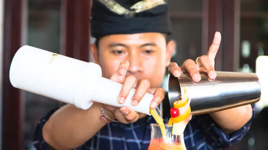 Bartender in traditional Balinese head wrap pouring a tropical cocktail into a glass with cherry and orange peel garnish.