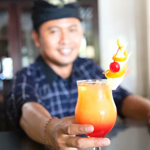 Refreshing tropical cocktail with vibrant orange and red layers, garnished with fruit, served by a smiling bartender in Bali.