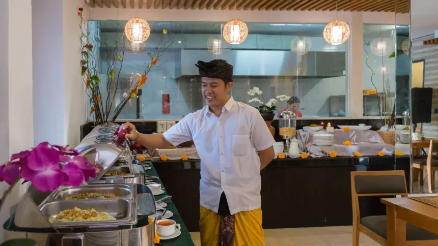 Smiling Balinese server in white shirt and yellow sarong arranging buffet trays at an open kitchen counter.