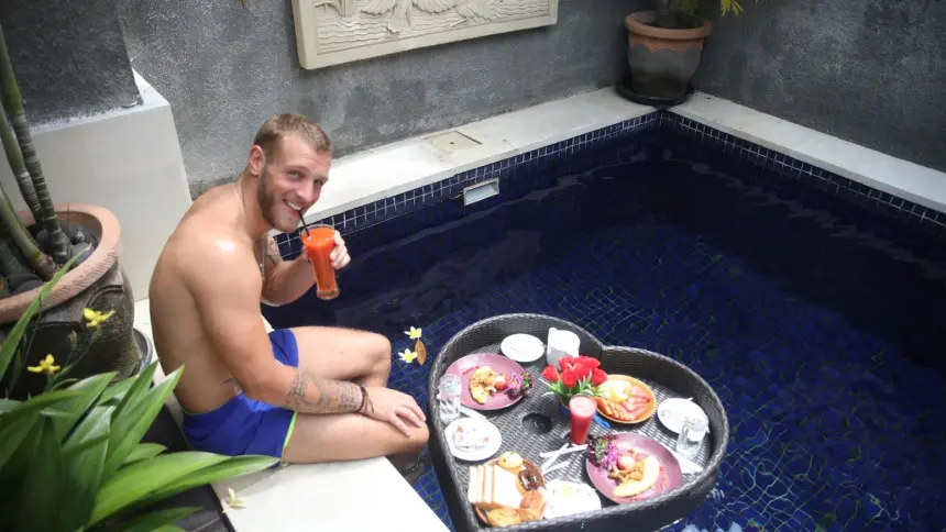 Man in swim shorts seated by a private pool with a heart-shaped wicker floating breakfast tray holding tropical fare.
