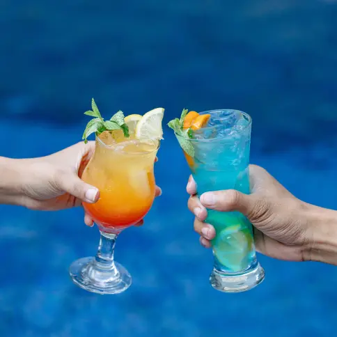 Two colorful cocktails with garnishes held over a vibrant blue pool background.