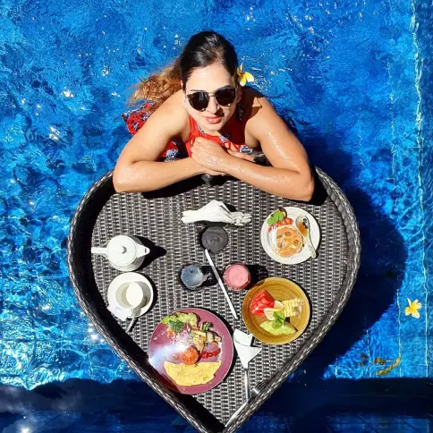 Overhead shot of a woman in red swimsuit posing in a pool behind a heart-shaped floating breakfast tray.