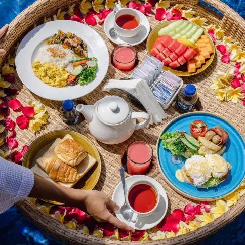 Floating breakfast tray with fruits, eggs Benedict, pastries, and tea in a pool surrounded by tropical flowers at a luxury villa.