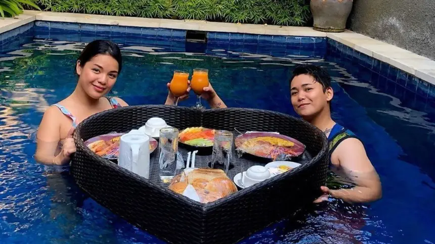 Two guests in a pool toasting with orange juice over a heart-shaped wicker floating breakfast tray.