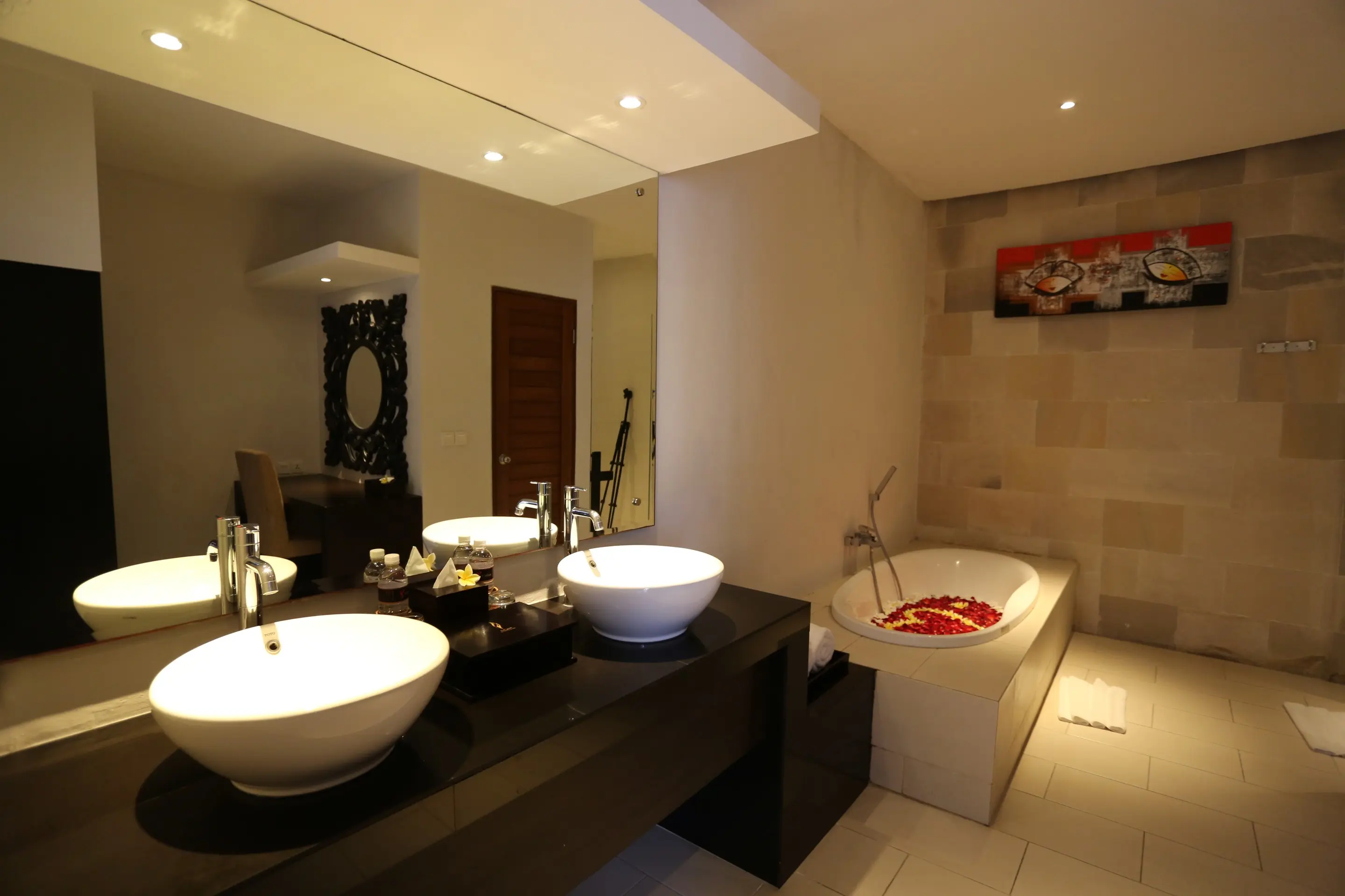 Spacious villa bathroom with twin vessel sinks, oversized mirror, freestanding bathtub with rose petals and warm stone walls.