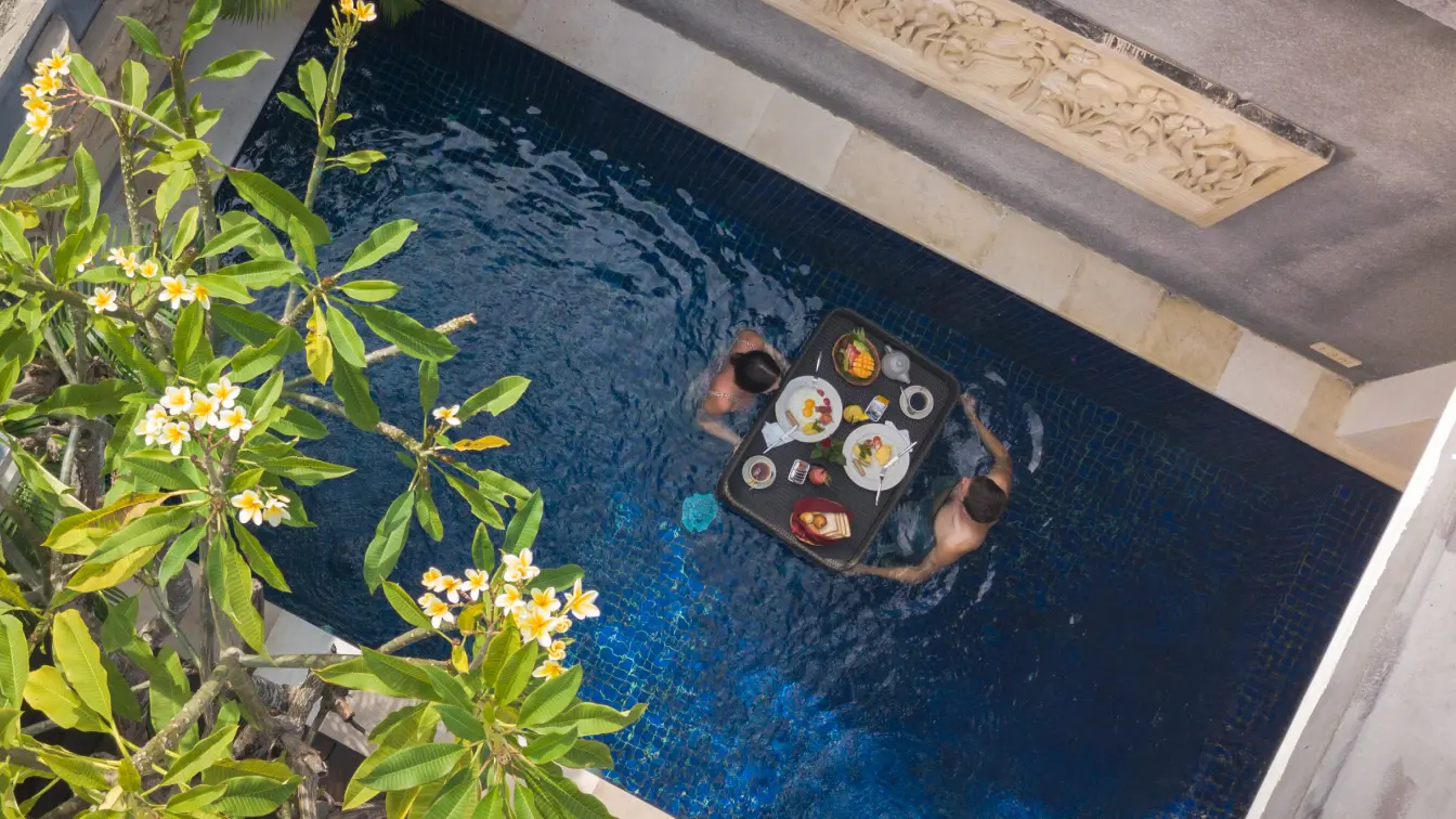 Overhead view of a couple in a private plunge pool with a black floating breakfast tray, framed by frangipani blossoms.