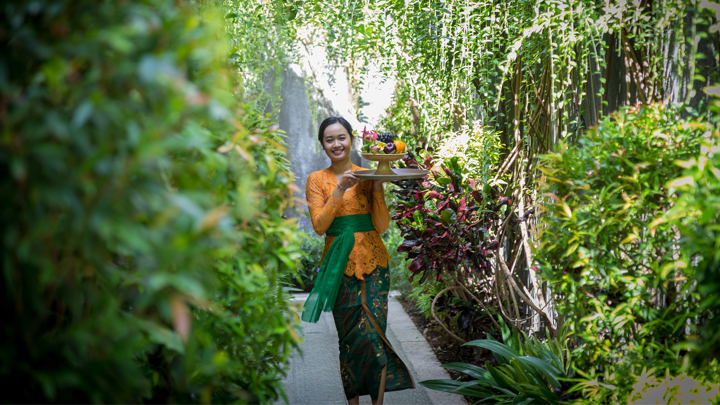 Balinese staff member in orange kebaya carrying a fruit tray along a leafy garden pathway at the villa.