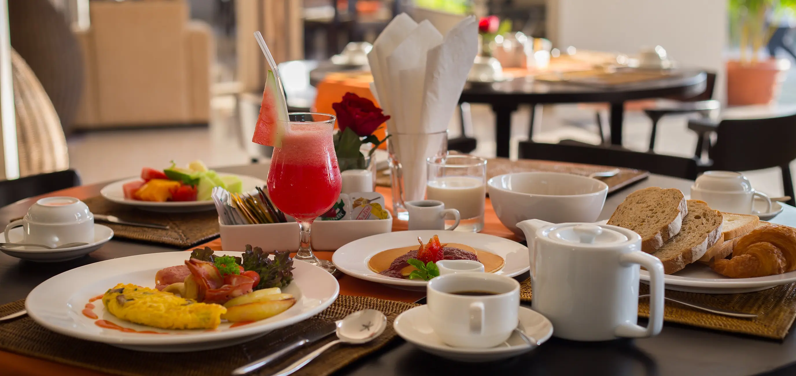 Indoor breakfast table set with an omelette plate, fresh fruit, a watermelon smoothie, pancakes, bread, and coffee.