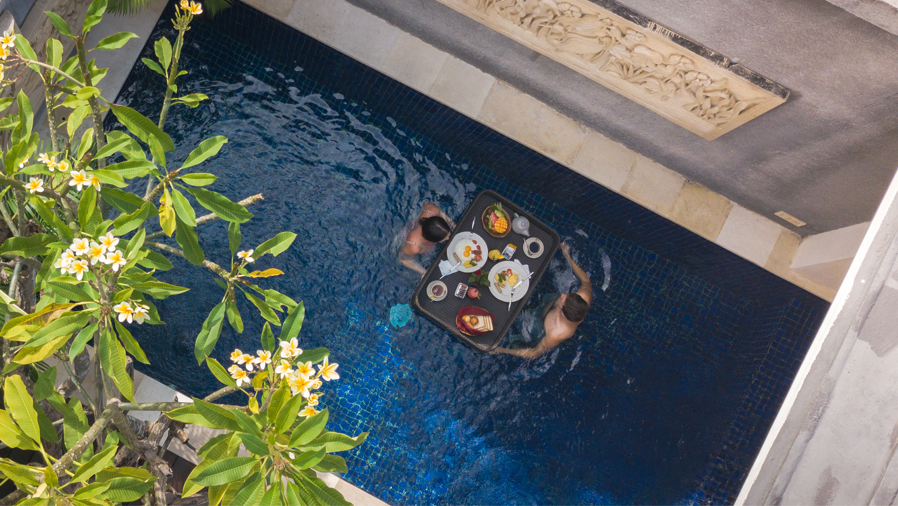 Overhead view of a couple in a private pool sharing a black floating breakfast tray surrounded by frangipani branches.