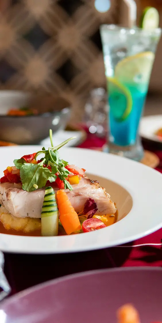 Gourmet fish dish with vibrant vegetables, paired with a tropical blue cocktail at The Light Exclusive Villas & Spa dining.