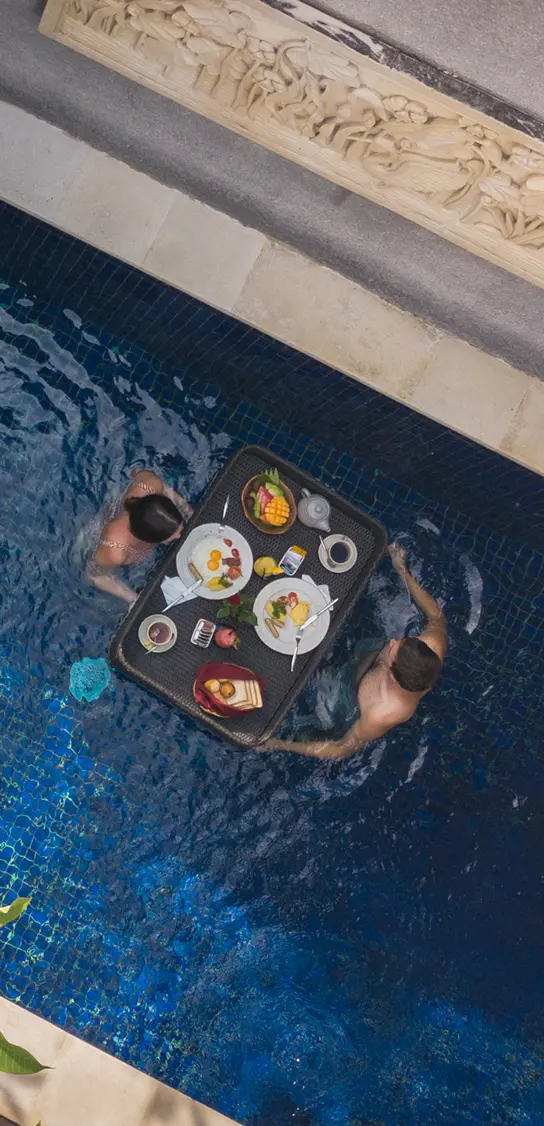 Couple in a private blue-tiled pool enjoying a wooden floating breakfast tray with tropical fruit and pastries.