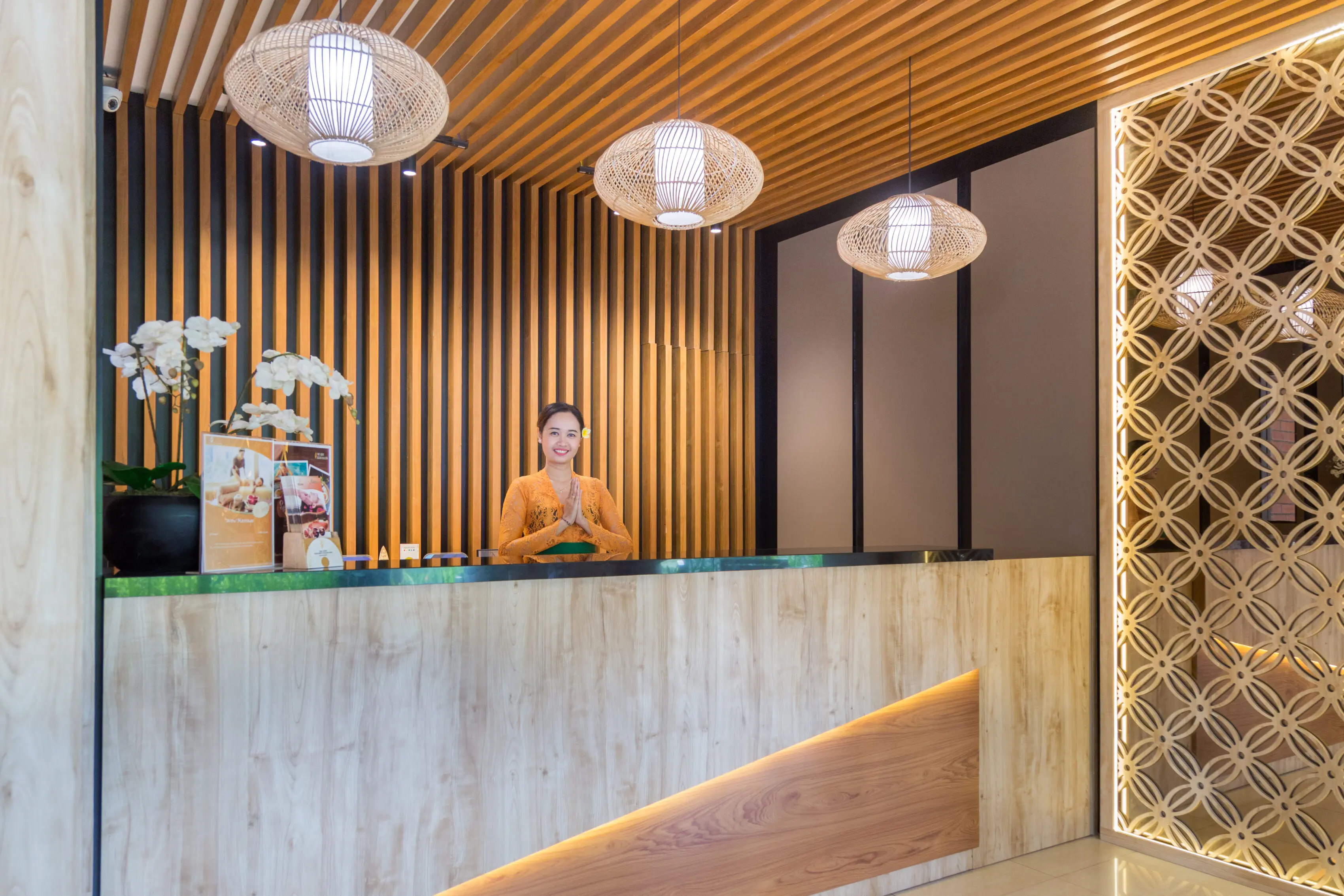 Balinese receptionist in orange kebaya greeting from behind a wooden reception desk under woven rattan pendant lights.