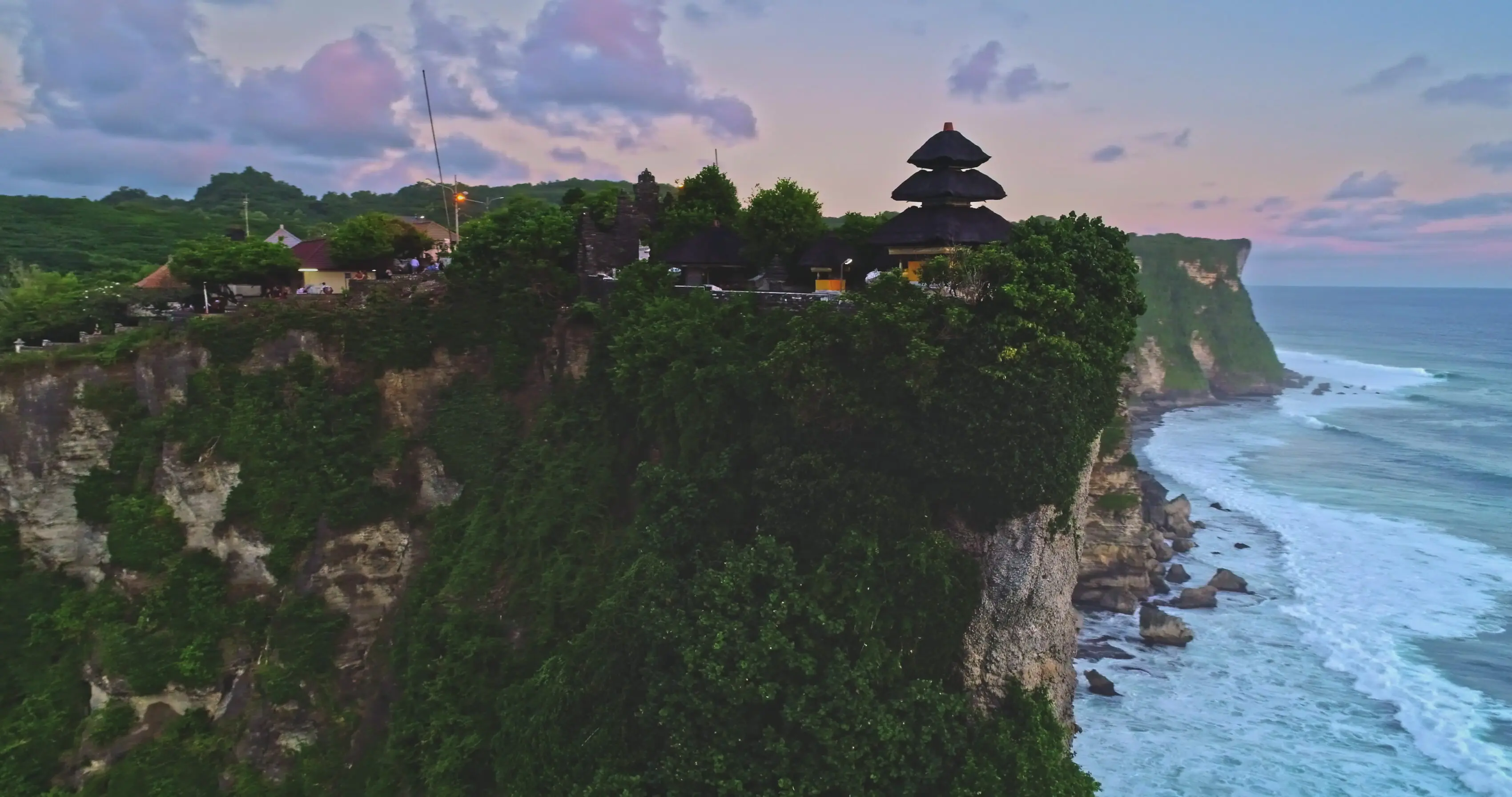 Aerial view of Uluwatu Temple perched on a cliffside overlooking the ocean at sunset.