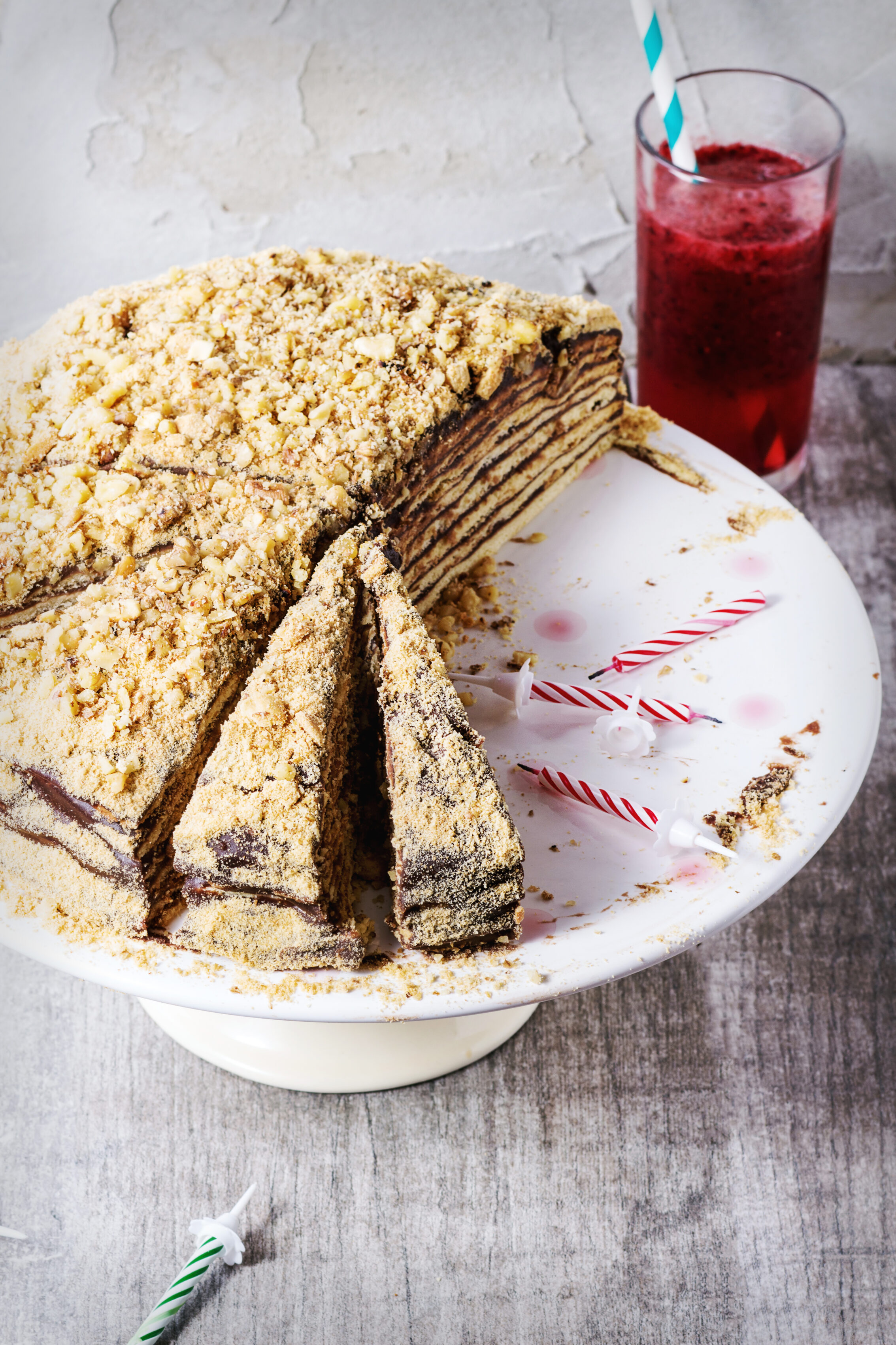 Sliced homemade Russian honey cake topped with crushed nuts beside a glass of berry smoothie and birthday candles.