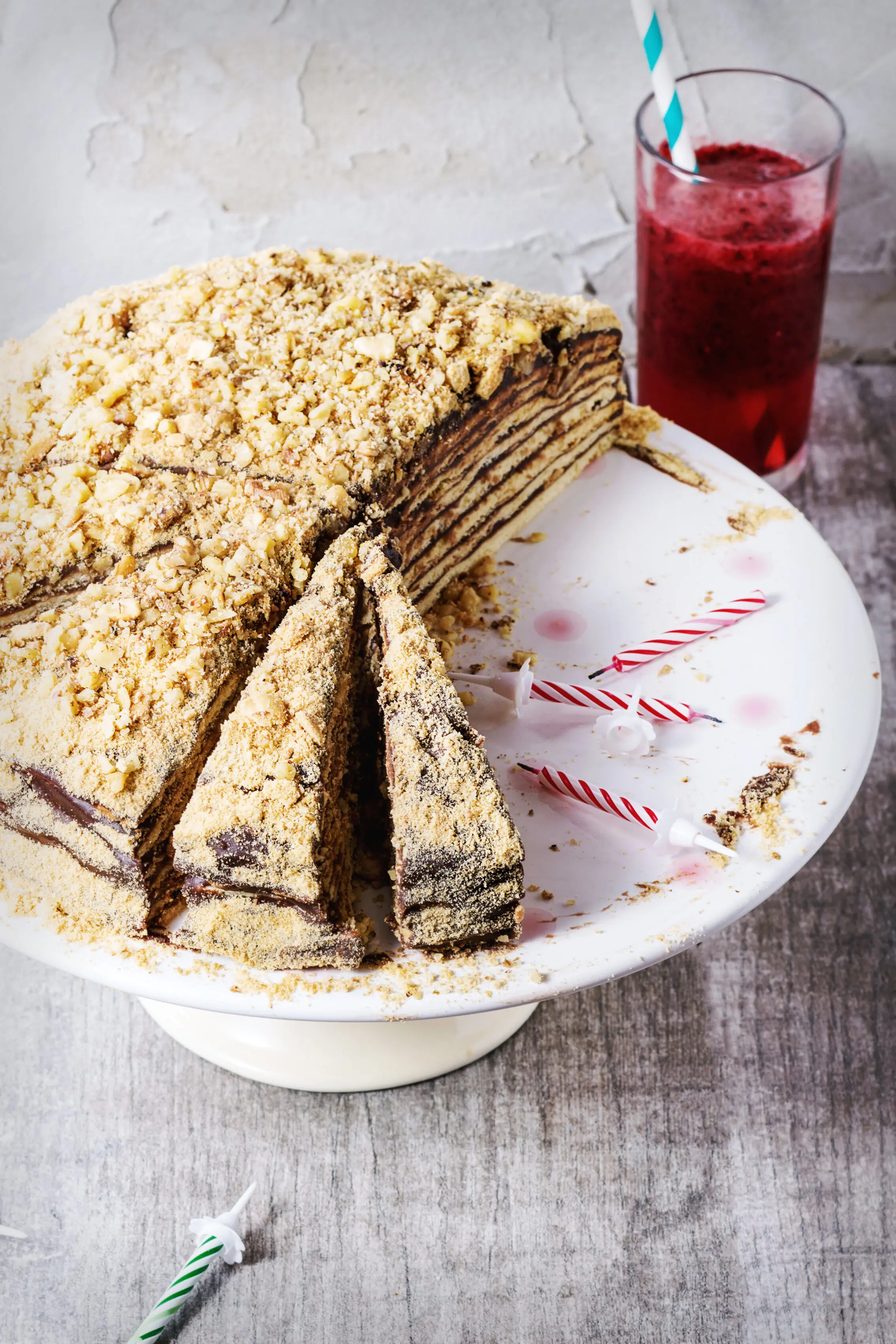 Sliced homemade Russian honey cake topped with crushed nuts beside a glass of berry smoothie and birthday candles.