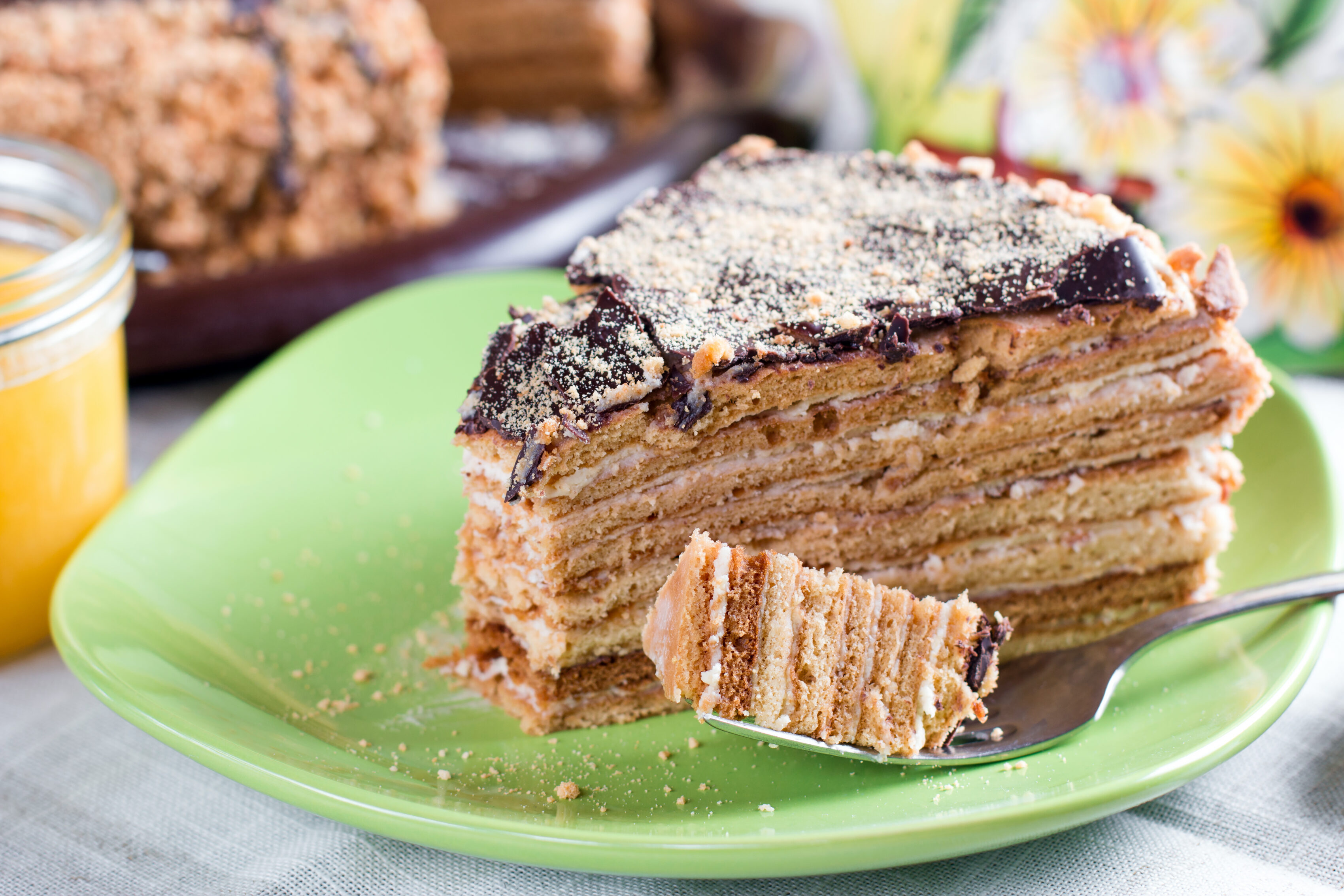 Slice of Russian Medovik honey cake with vanilla cream, chocolate glaze and crumb topping on a green plate.