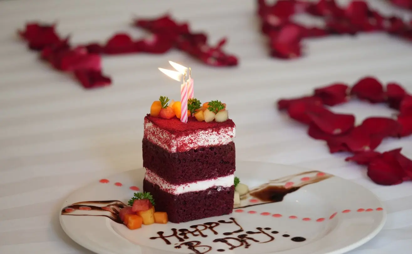 Red velvet cake with candles on an elegant plate, surrounded by rose petals, for a surprise birthday celebration.