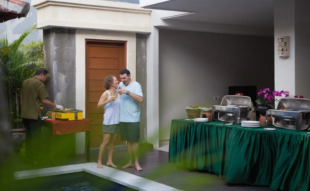 Couple toasting drinks beside a poolside BBQ buffet with green-skirted tables and chafing dishes at the villa.