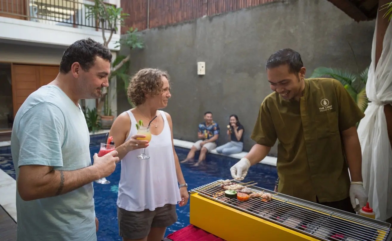 Guests with cocktails chatting with a smiling Balinese chef grilling satay skewers on a yellow BBQ beside a private pool.