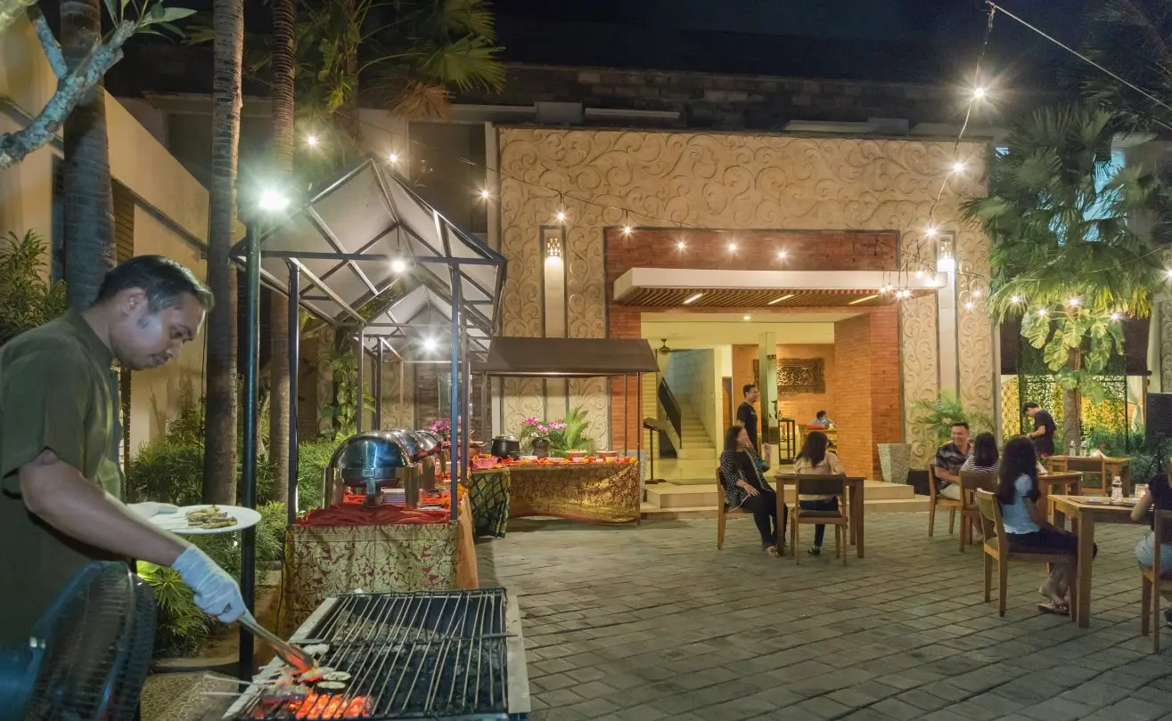 Chef grilling satay over a charcoal BBQ at an open-air dinner buffet under string lights in the villa courtyard.