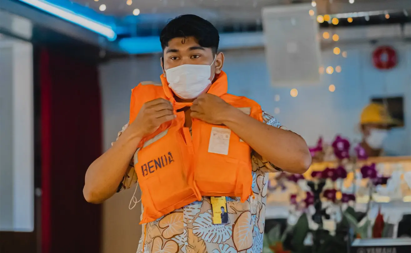 Person in floral shirt adjusting life jacket on a cruise ship, surrounded by twinkling lights and orchids.