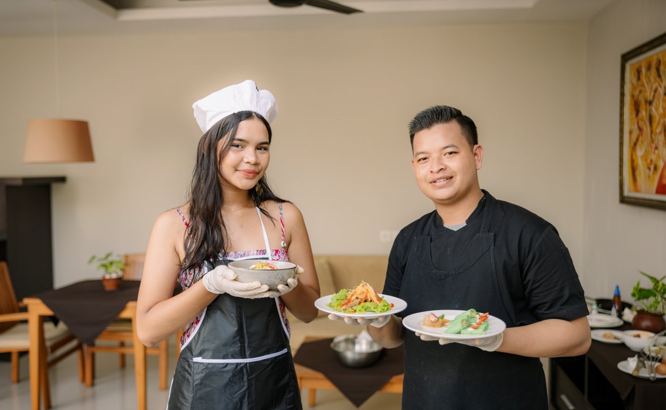 Smiling guest in chef's hat and a chef in black apron holding plates of finished dishes inside the villa living area.