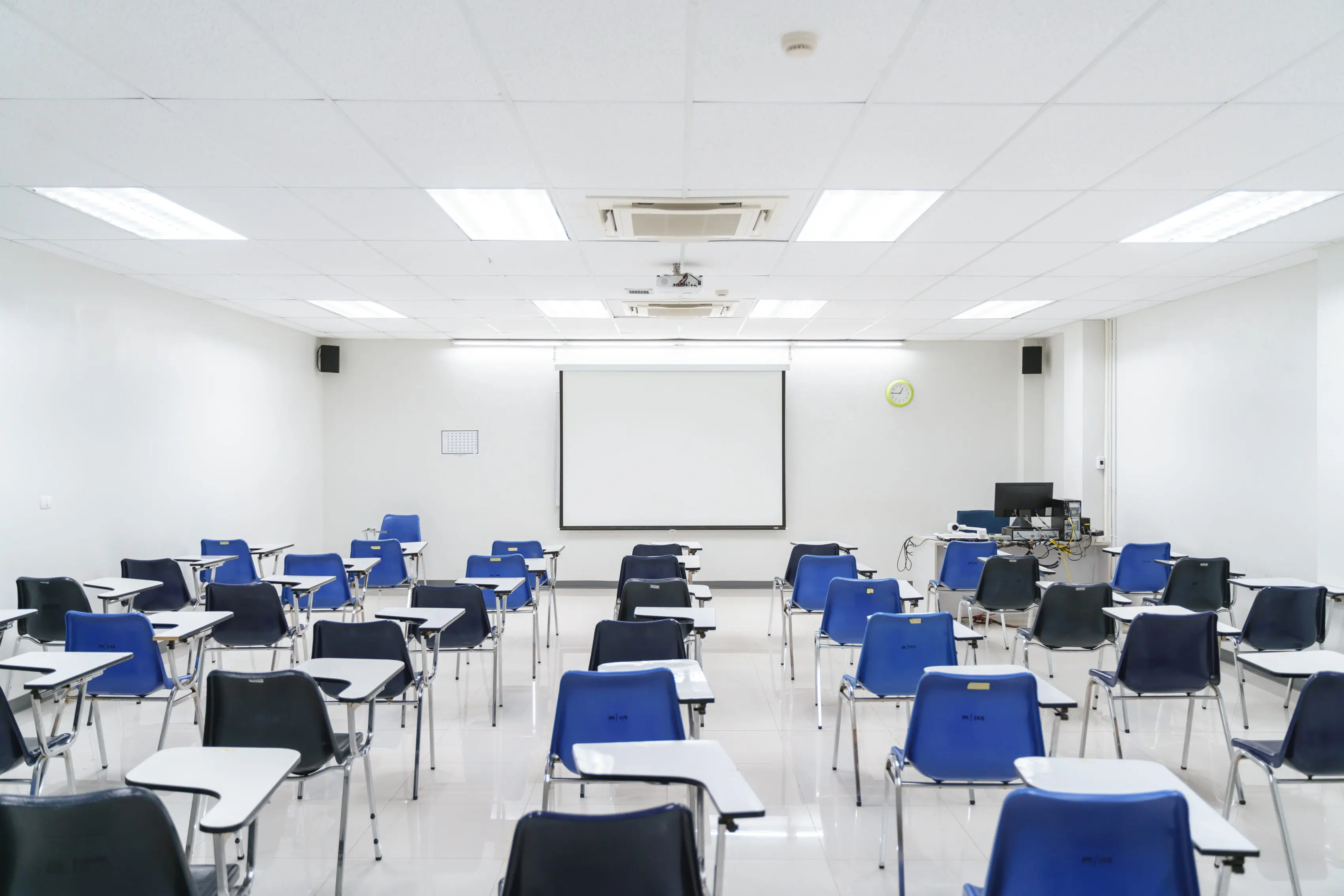 Empty conference room with rows of blue tablet-arm chairs facing a projector screen, ready for a meeting.