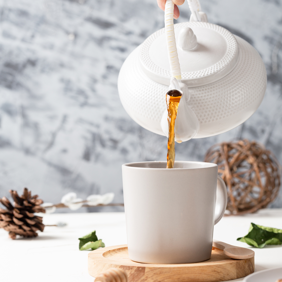 White textured teapot pouring tea into a mug on a wooden tray beside a pinecone, on a grey marbled background.