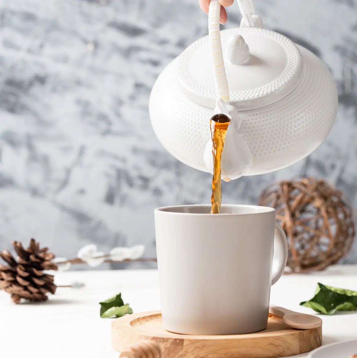White textured teapot pouring tea into a mug on a wooden tray beside a pinecone, on a grey marbled background.