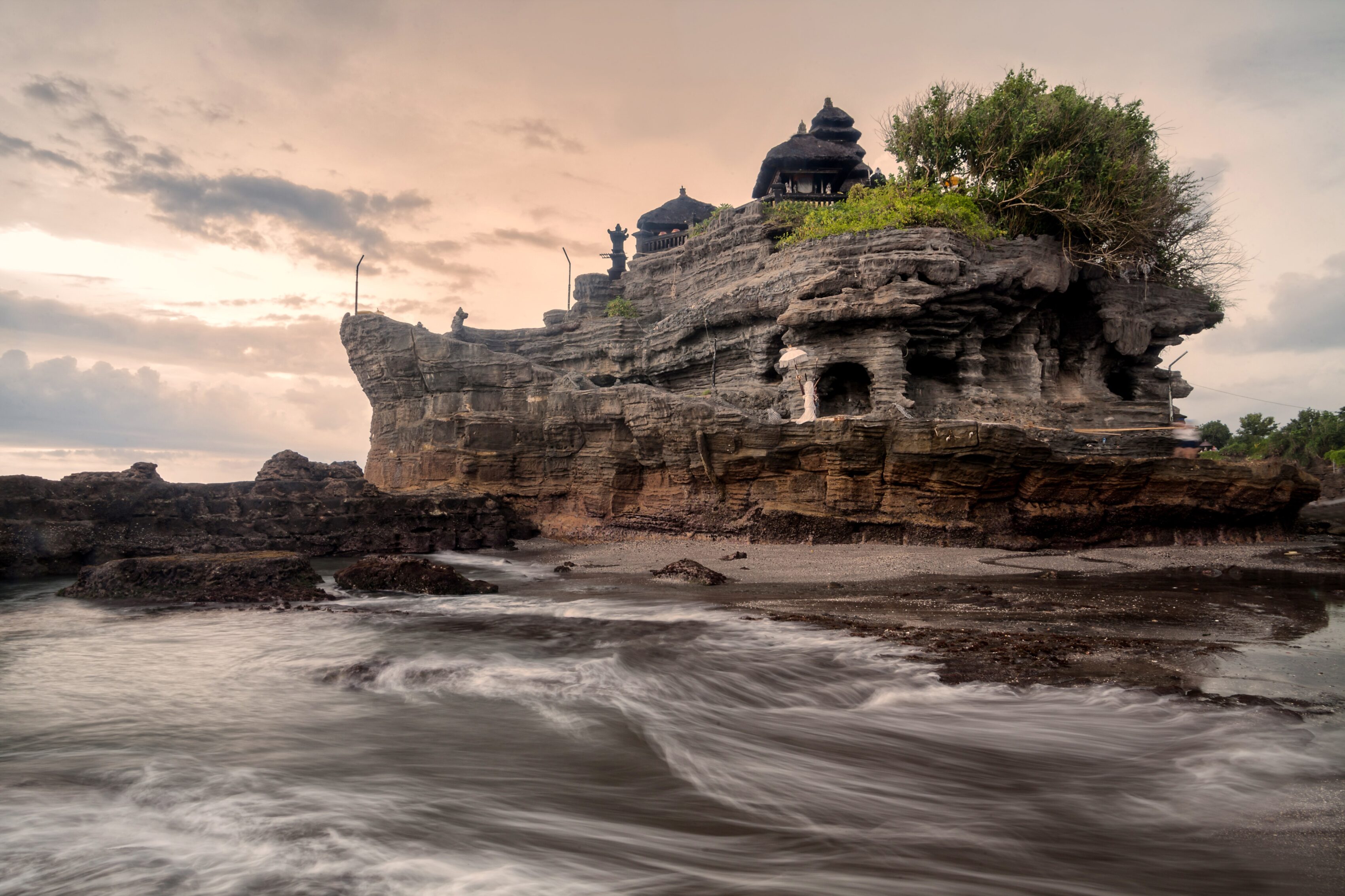 Tanah Lot sea temple on its weathered rock formation with waves rolling onto the shore under a dusky sky.