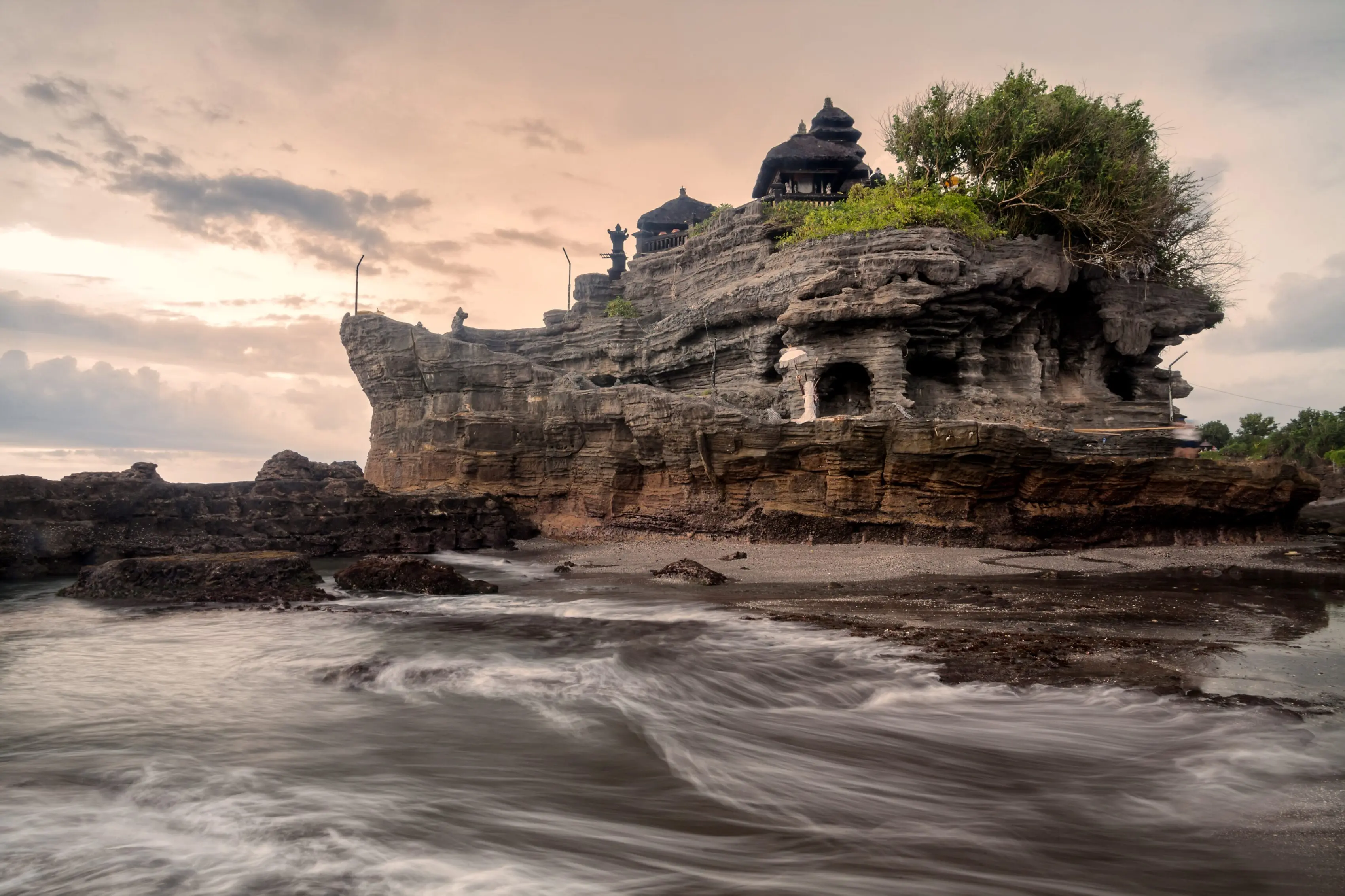Tanah Lot sea temple on its weathered rock formation with waves rolling onto the shore under a dusky sky.