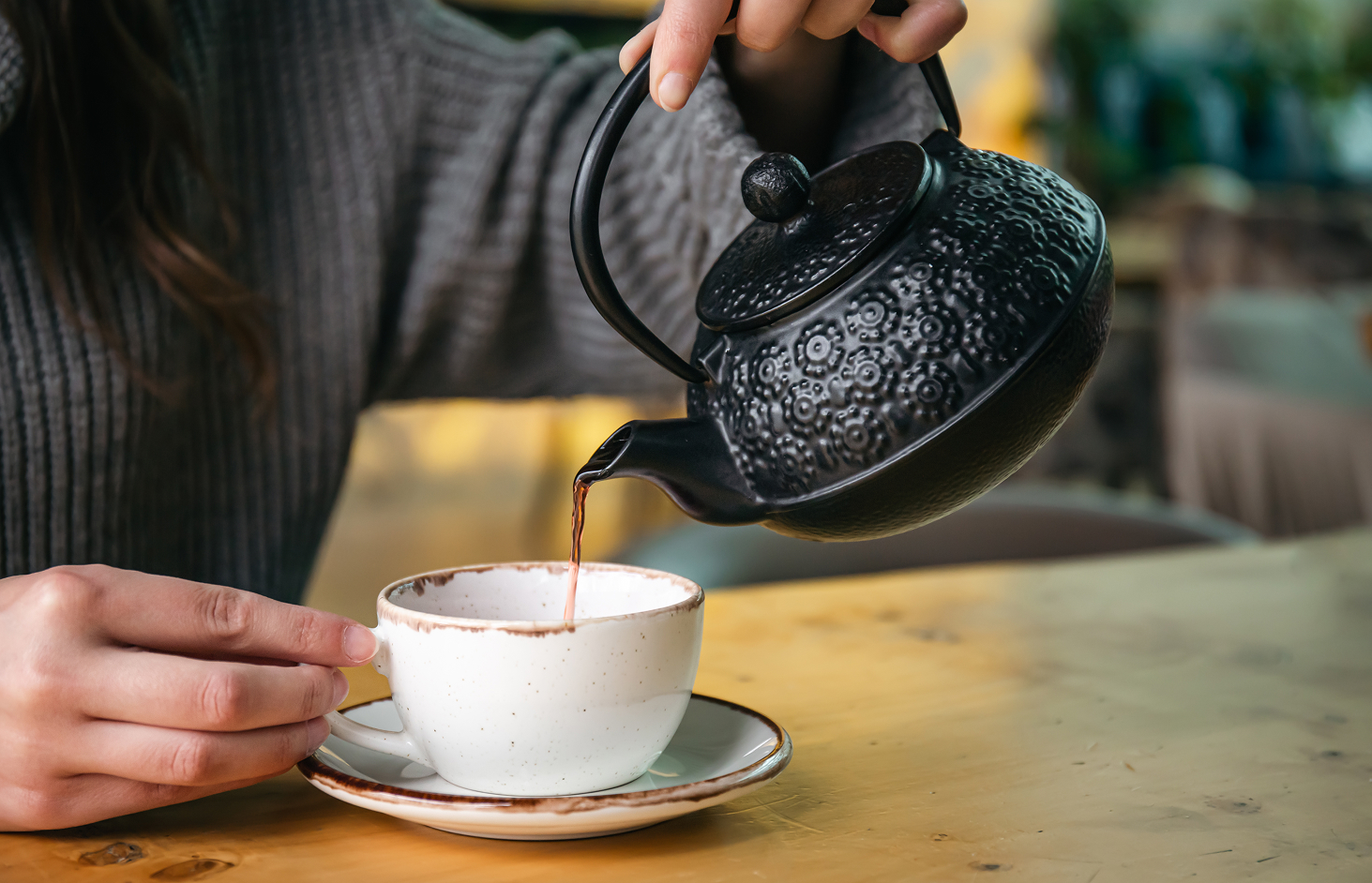 Woman pouring tea from a textured black cast-iron teapot into a ceramic cup on a wooden café table.